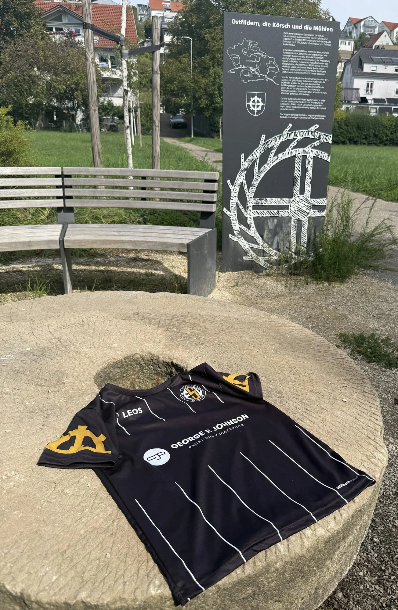 Trikot der FSG Ostfildern auf einem alten Mühlstein, dahinter eine Tafel über die Körsch und die Mühlen Ostfilderns